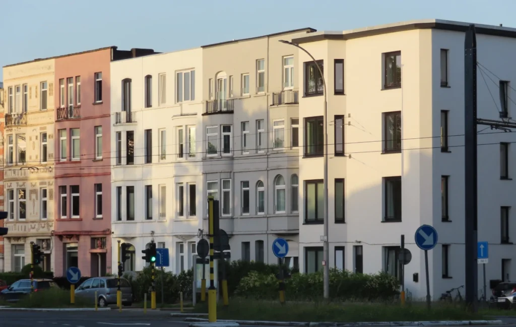 Een rij van aaneengesloten stadsgebouwen (rijhuizen of appartementsgebouwen) aan een stedelijk kruispunt, gefotografeerd in warm avondlicht. De gebouwen zijn drie à vier verdiepingen hoog en hebben een gevarieerde gevel: van links naar rechts gaan de kleuren van roze over cremewit naar lichtgrijs en helder wit. Het meest linkse gebouw heeft decoratieve stucornamenten in art-nouveaustijl. Op de voorgrond zijn verkeerstekens zichtbaar — blauwe rondebordjes met een witte pijl, een groen verkeerslicht, en gele paaltjes langs het voetpad. Er staan enkele geparkeerde auto's en er is lage beplanting zichtbaar tussen de rijweg en de gebouwen. De lucht is lichtblauw met een zachte gloed, wat wijst op vroege ochtend of late avond.